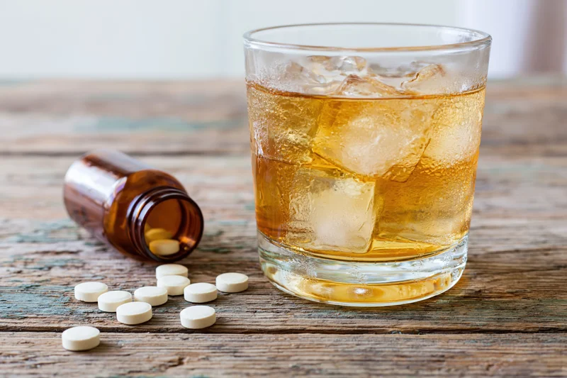 Glass of amber whiskey with ice beside spilled pills on a wooden table