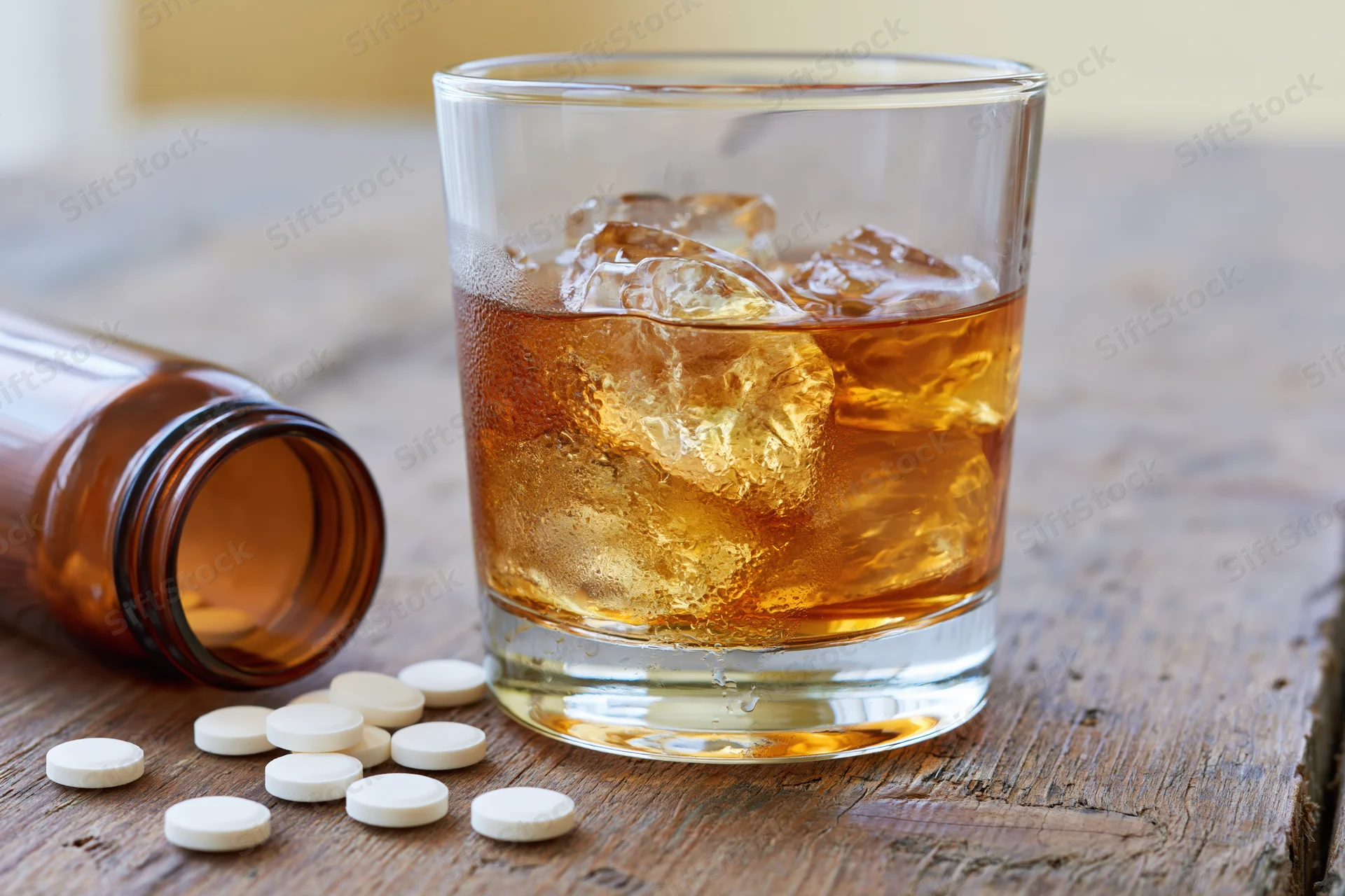 Whiskey in a rocks glass with ice beside scattered pills and a bottle on a wooden table