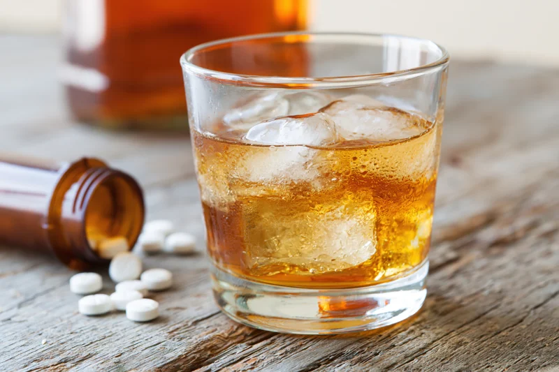 Whiskey in a rocks glass with ice on a wooden table beside a bottle and scattered pills