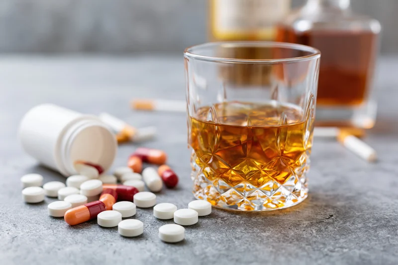 Whiskey in a rocks glass beside scattered pills and cigarettes on a grey concrete surface