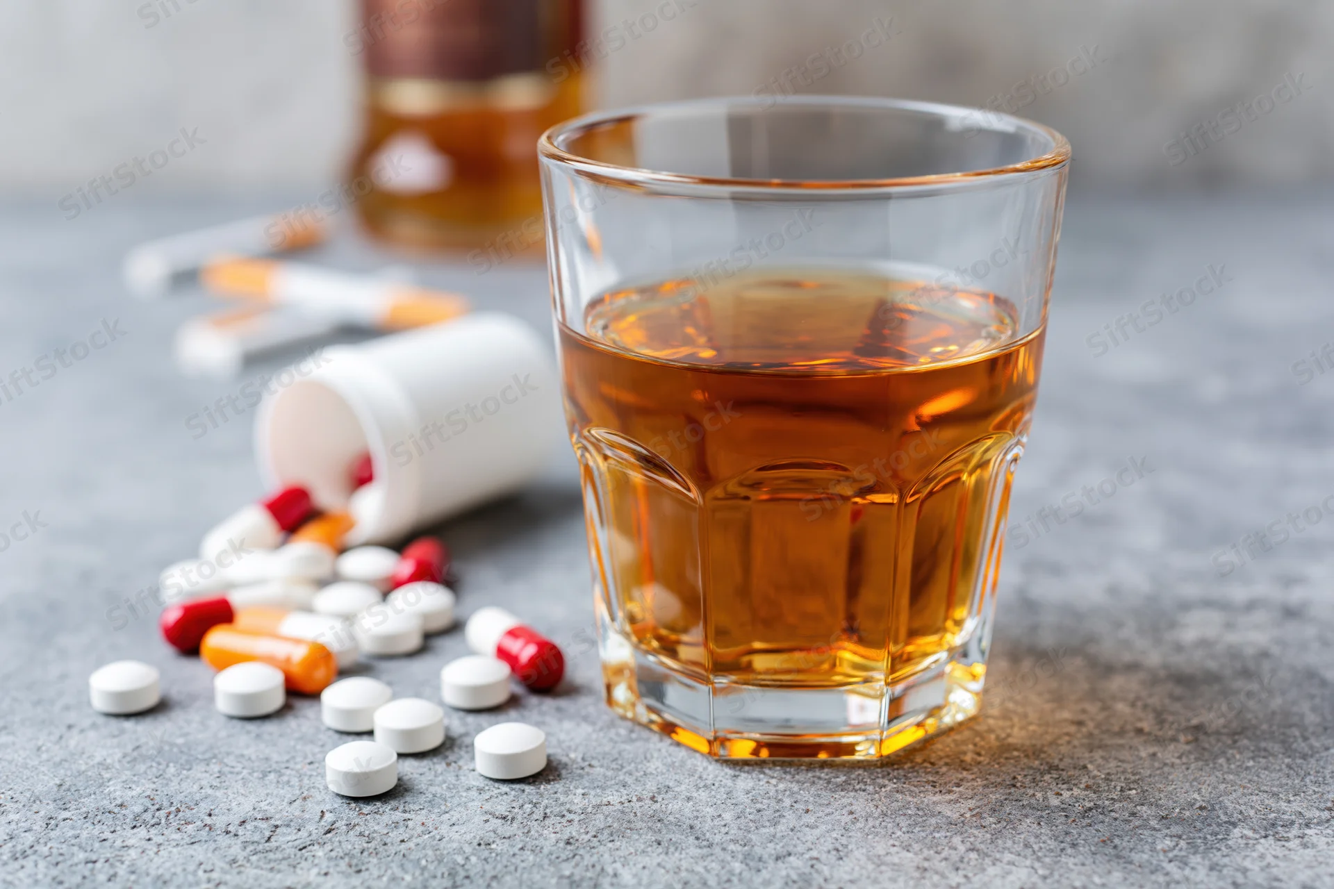 Glass of amber whiskey on grey surface with scattered pills and bottle in background