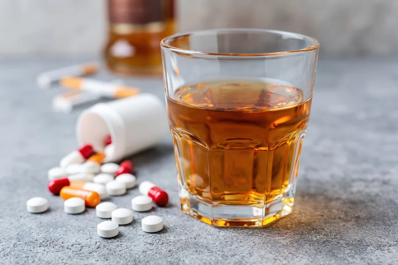 Glass of amber whiskey on grey surface with scattered pills and bottle in background