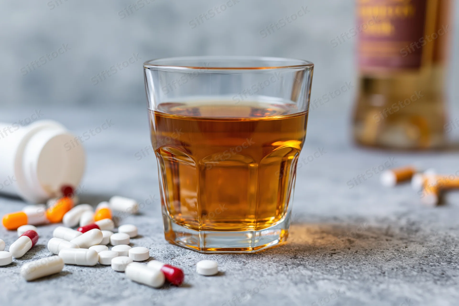 Glass of amber whiskey on grey concrete surface surrounded by scattered pills and cigarettes