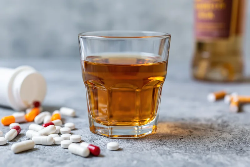 Glass of amber whiskey on grey concrete surface surrounded by scattered pills and cigarettes