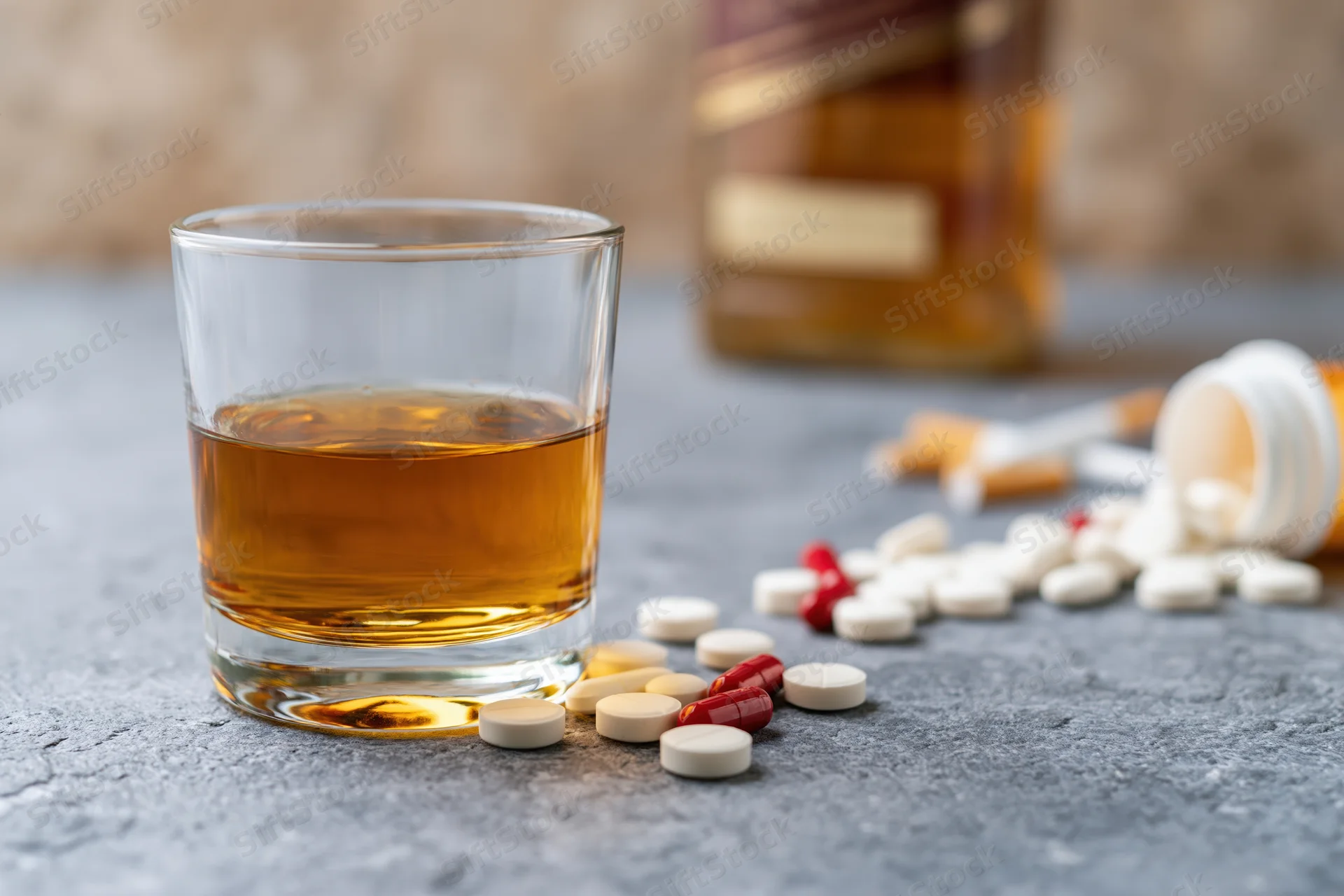 Whiskey glass with scattered pills and bottle on grey countertop
