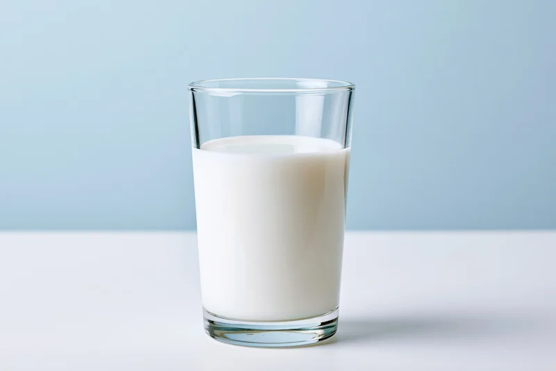 A clear glass filled with white milk sitting on a white surface against a light blue background