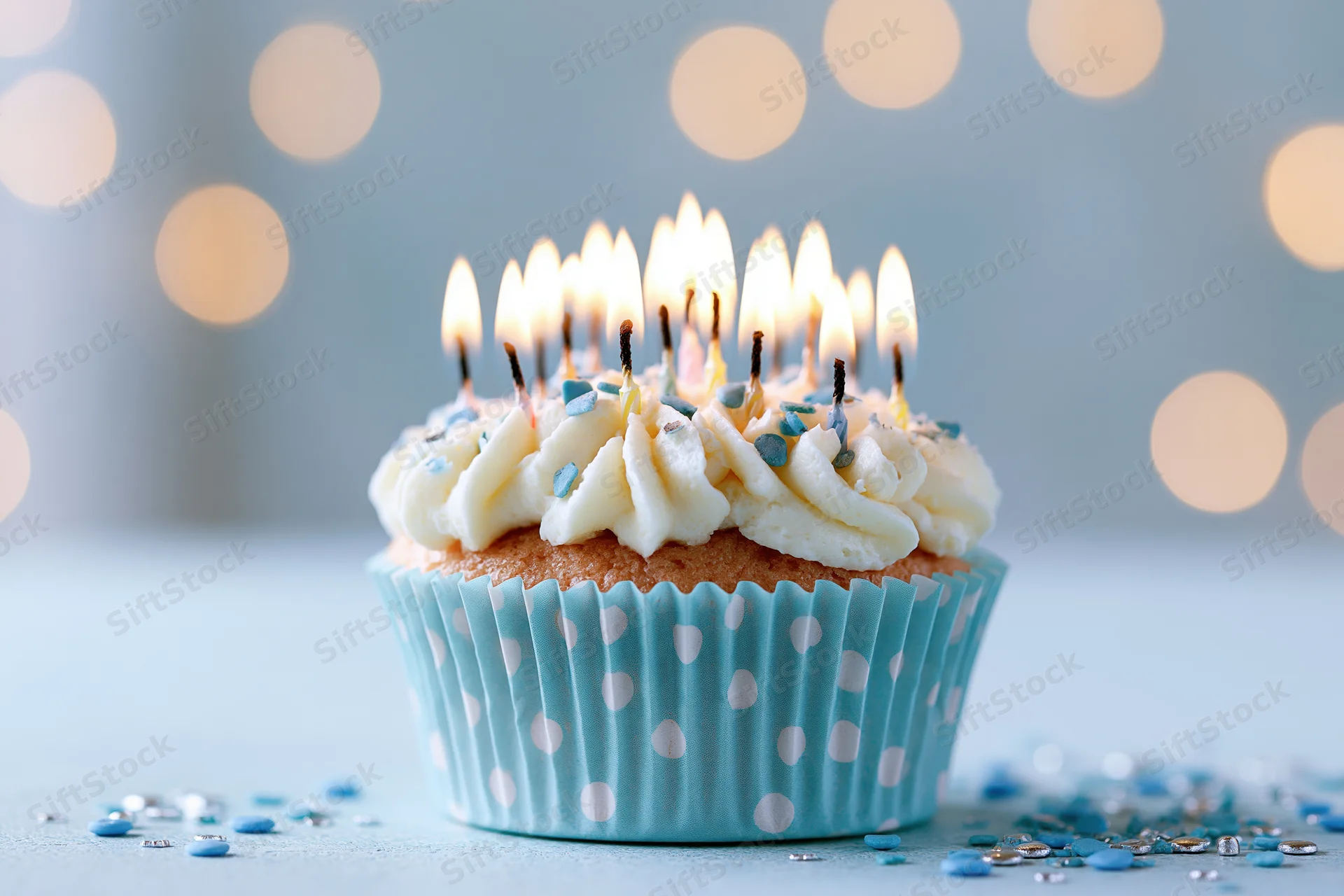 Single birthday cupcake with lit candles in blue polka-dot wrapper on light surface with bokeh lights behind