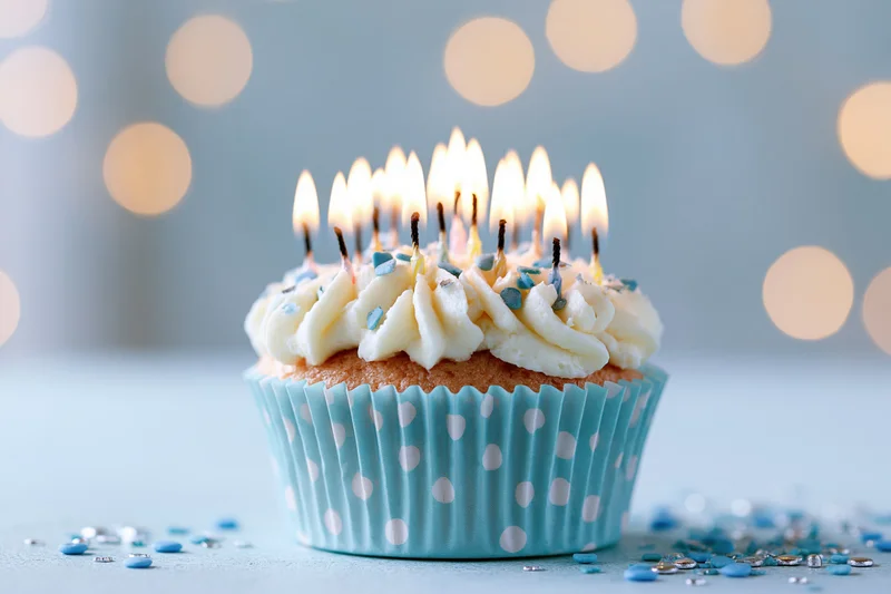 Single birthday cupcake with lit candles in blue polka-dot wrapper on light surface with bokeh lights behind