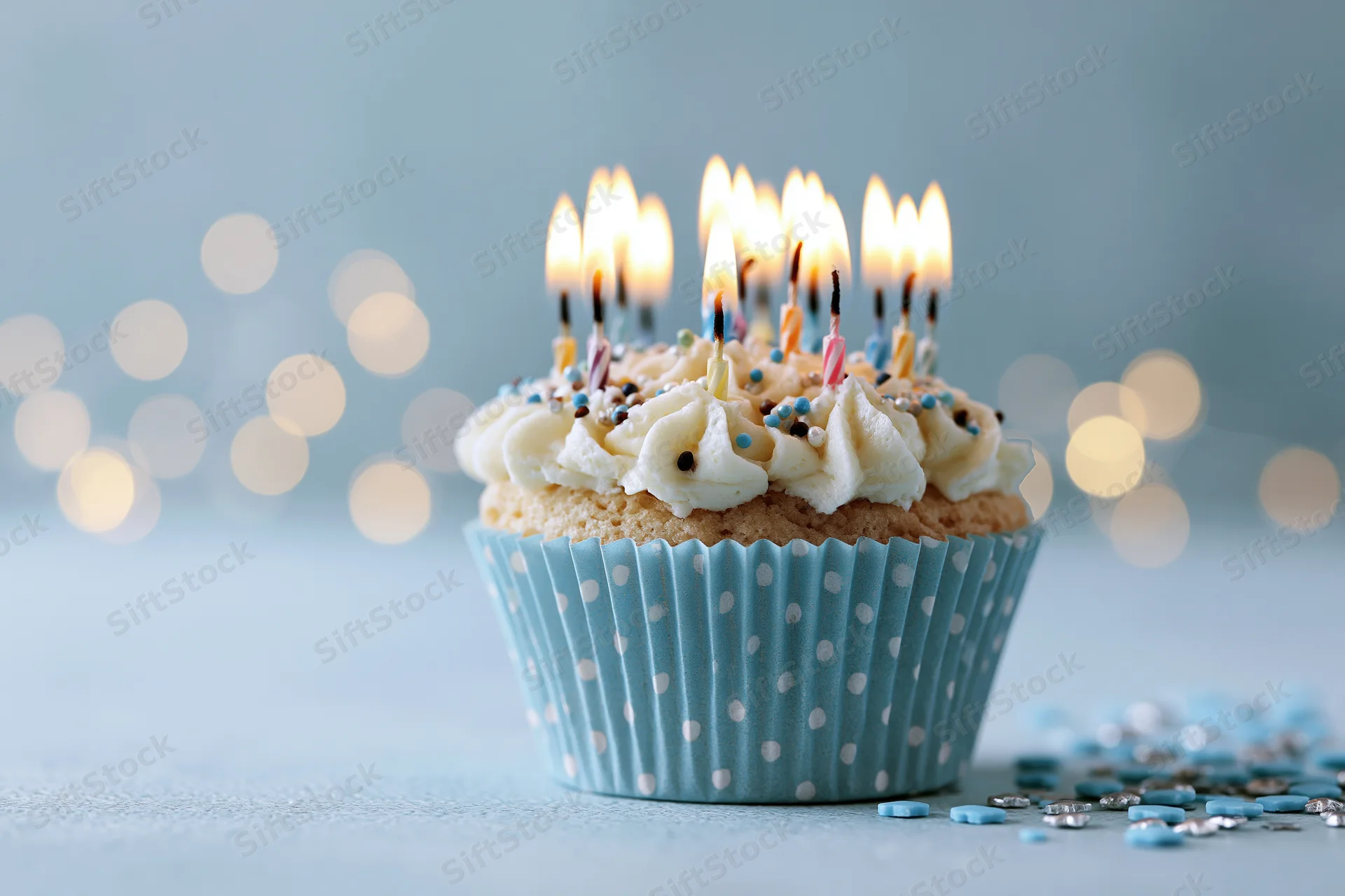Single birthday cupcake with lit candles in blue polka-dot liner on light surface with bokeh background