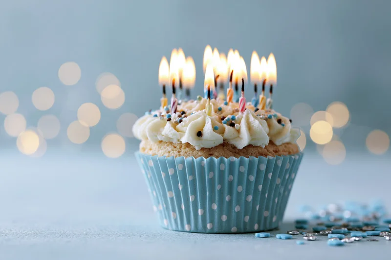 Single birthday cupcake with lit candles in blue polka-dot liner on light surface with bokeh background
