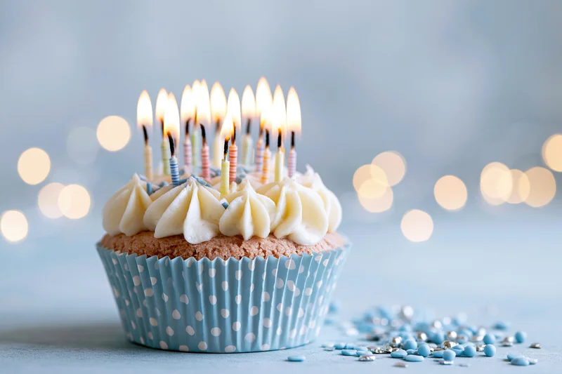 Single birthday cupcake with lit candles in blue polka-dot wrapper on light surface with scattered sprinkles and bokeh lights behind