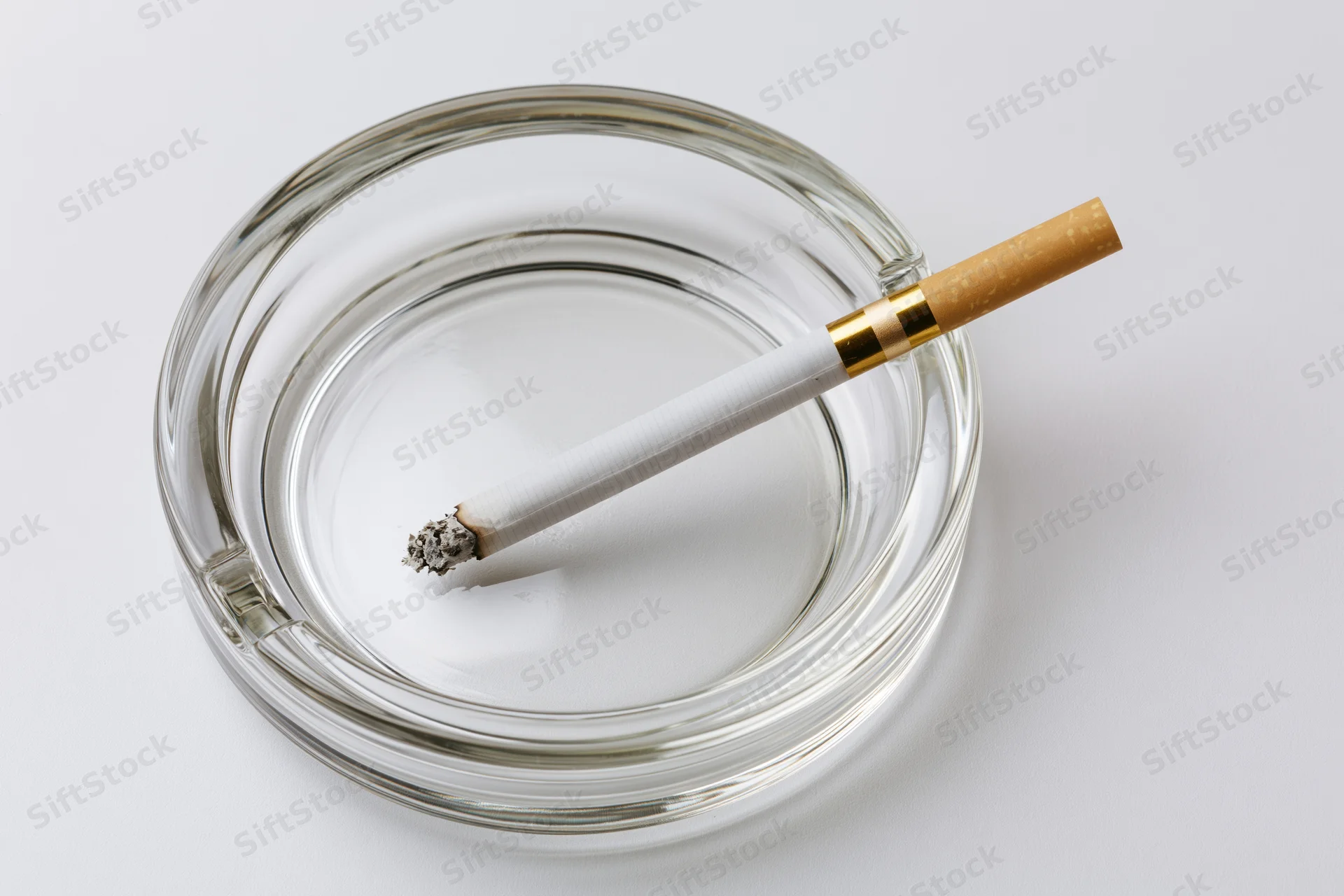 Lit cigarette resting in a clear glass ashtray on white surface