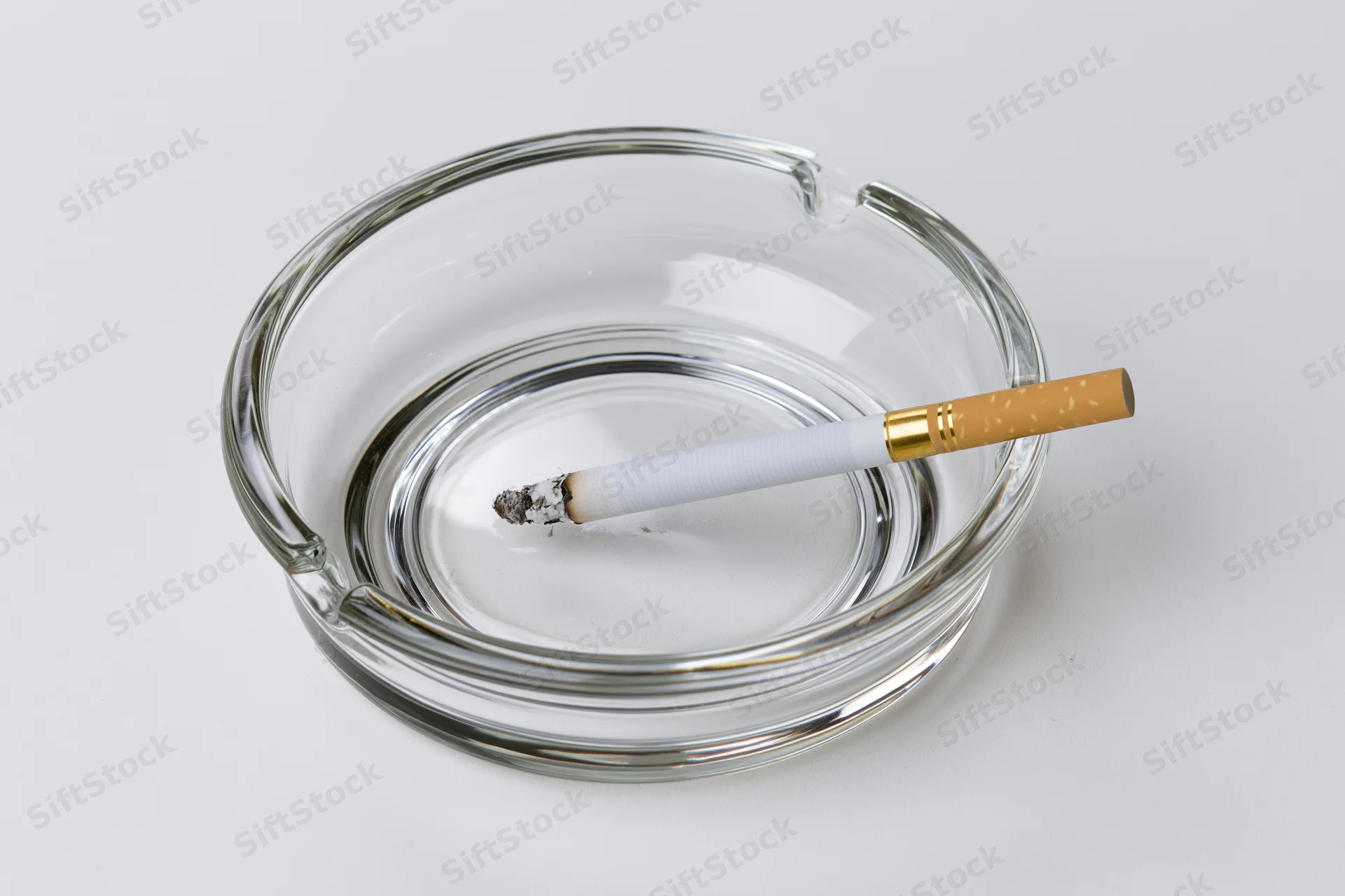 Lit cigarette resting in a clear glass ashtray on white background