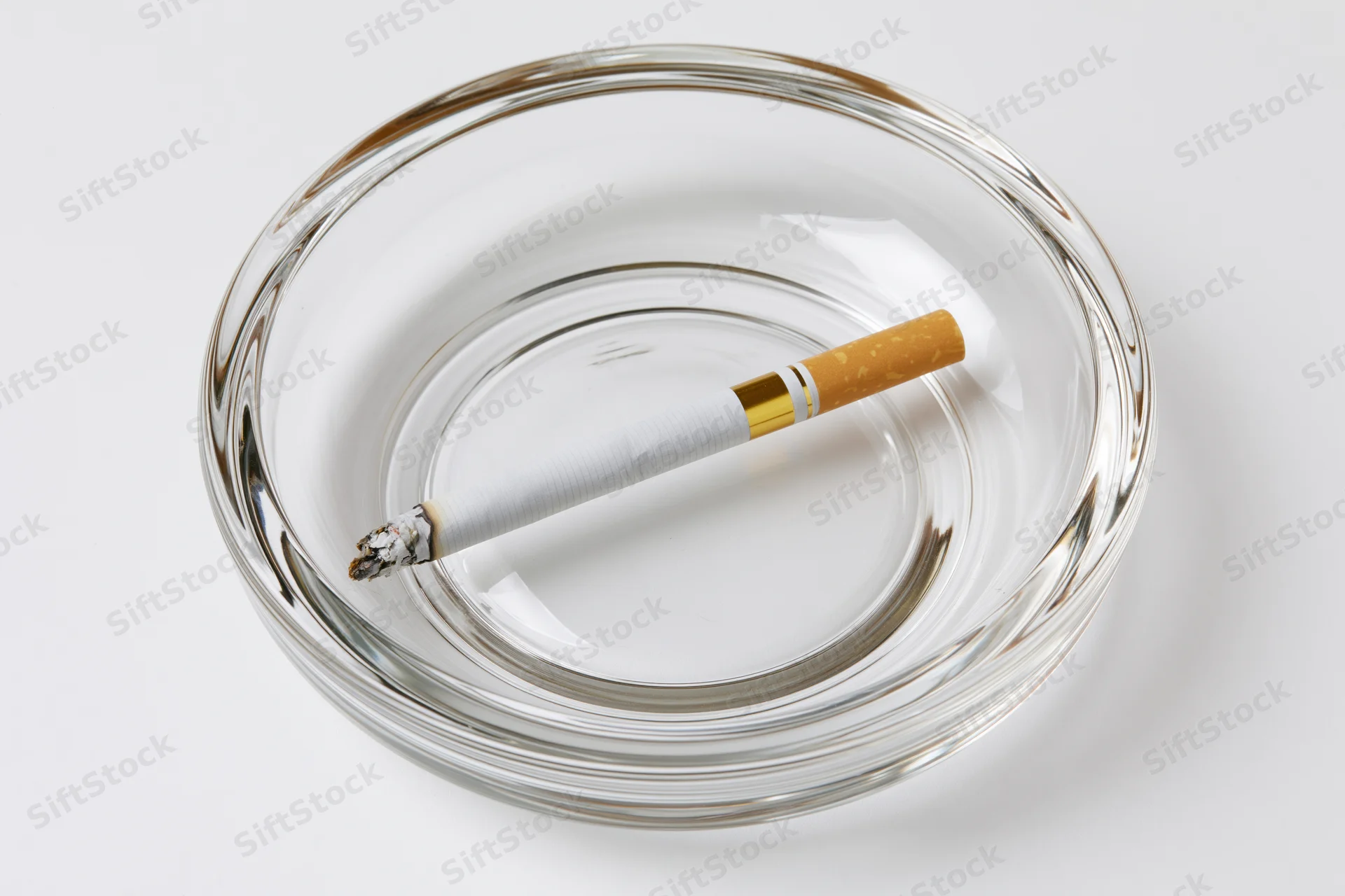 Lit cigarette resting in a clear glass ashtray on white background