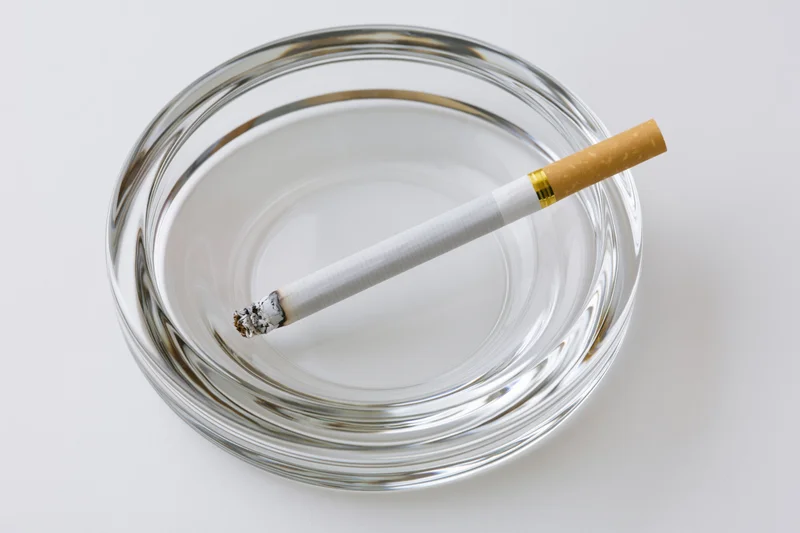 Lit cigarette resting in a clear glass ashtray on white surface