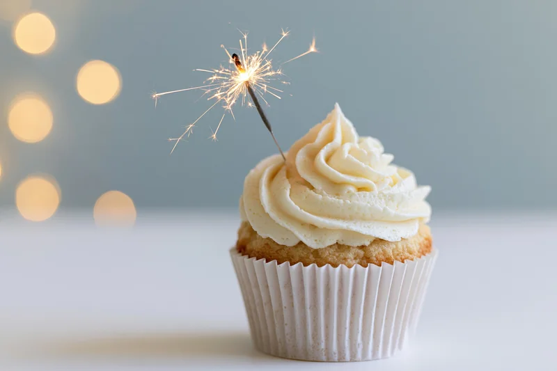 Vanilla cupcake with swirled white buttercream frosting and lit sparkler on white surface with bokeh lights in background
