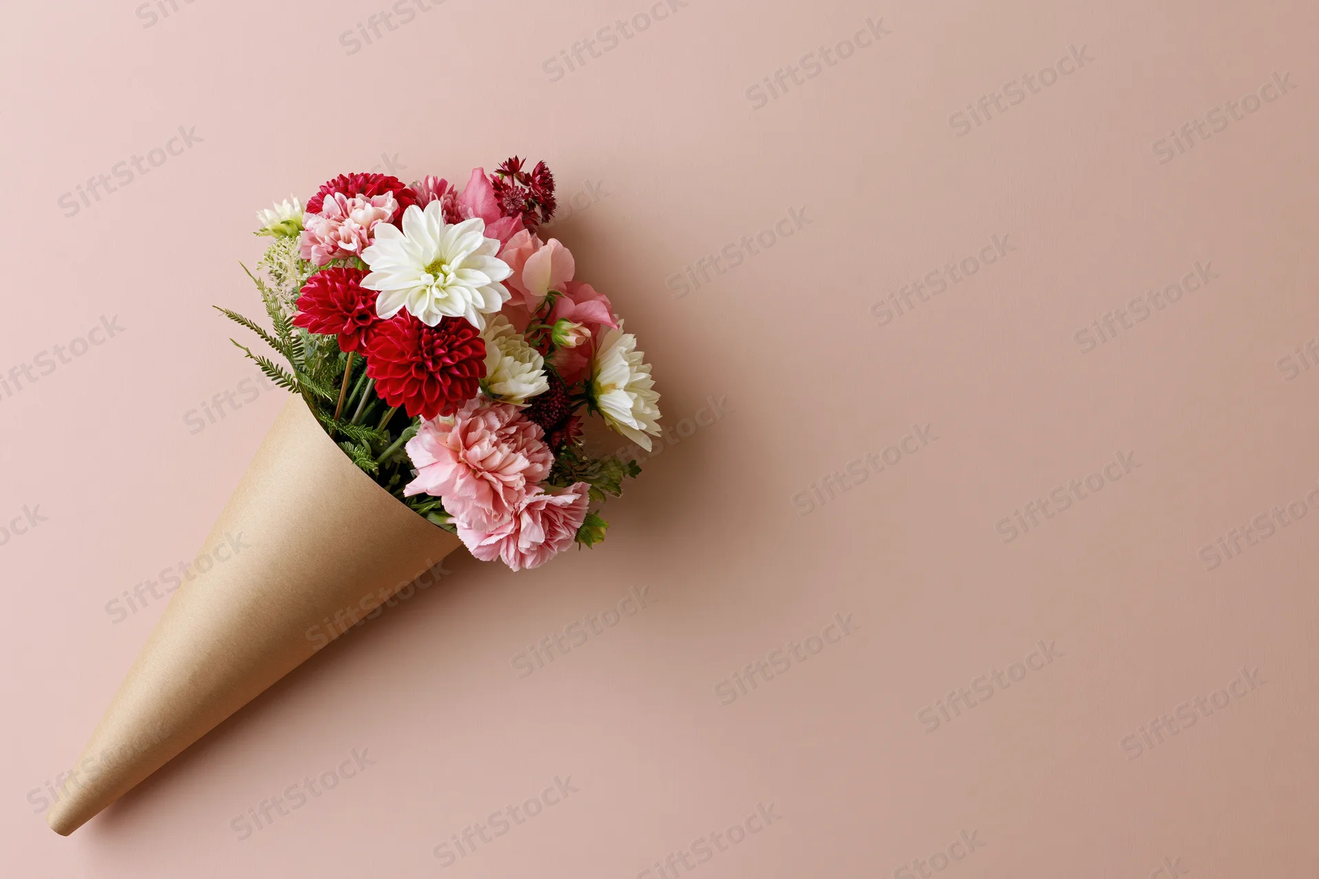 Small bouquet of mixed red white and pink flowers wrapped in brown paper cone on pastel pink background