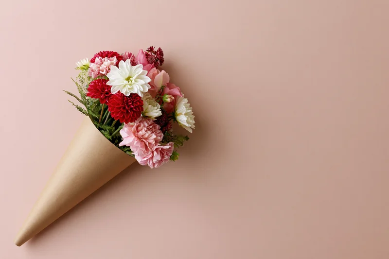 Small bouquet of mixed red white and pink flowers wrapped in brown paper cone on pastel pink background