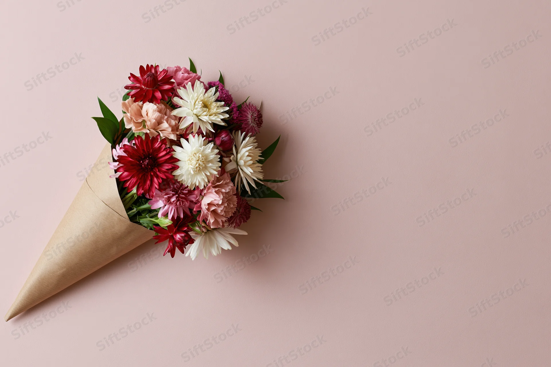 Small bouquet of mixed red white and pink flowers wrapped in brown paper cone on pastel pink background