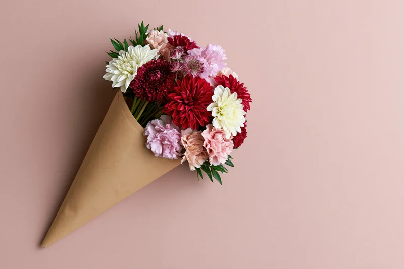 Small bouquet of mixed red white and pink flowers wrapped in brown paper cone on pastel pink background