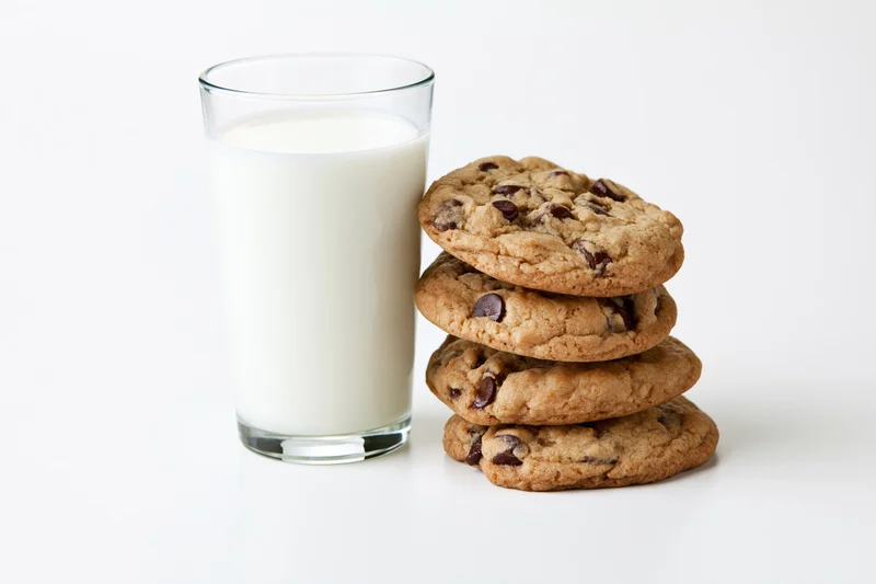 Stack of chocolate chip cookies beside a full glass of milk on white surface
