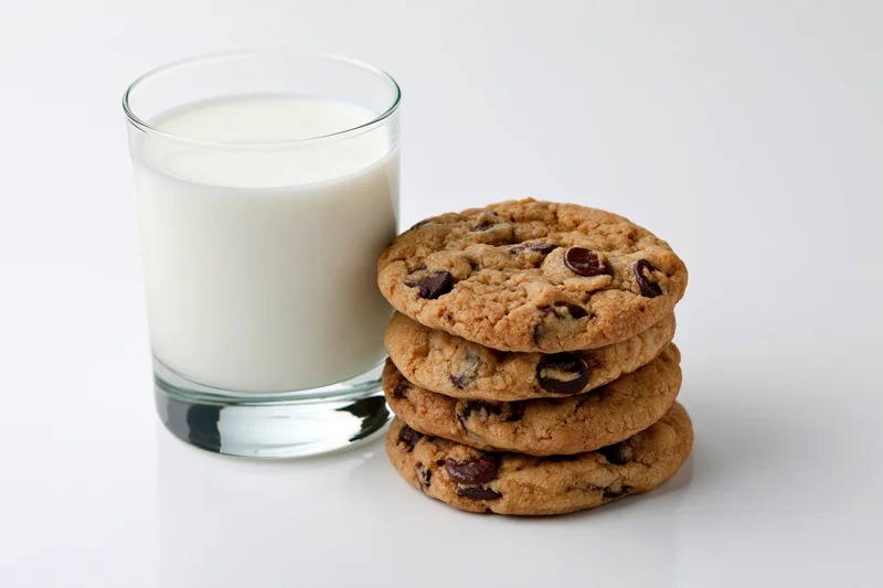 Stack of four chocolate chip cookies beside a full glass of milk on white surface