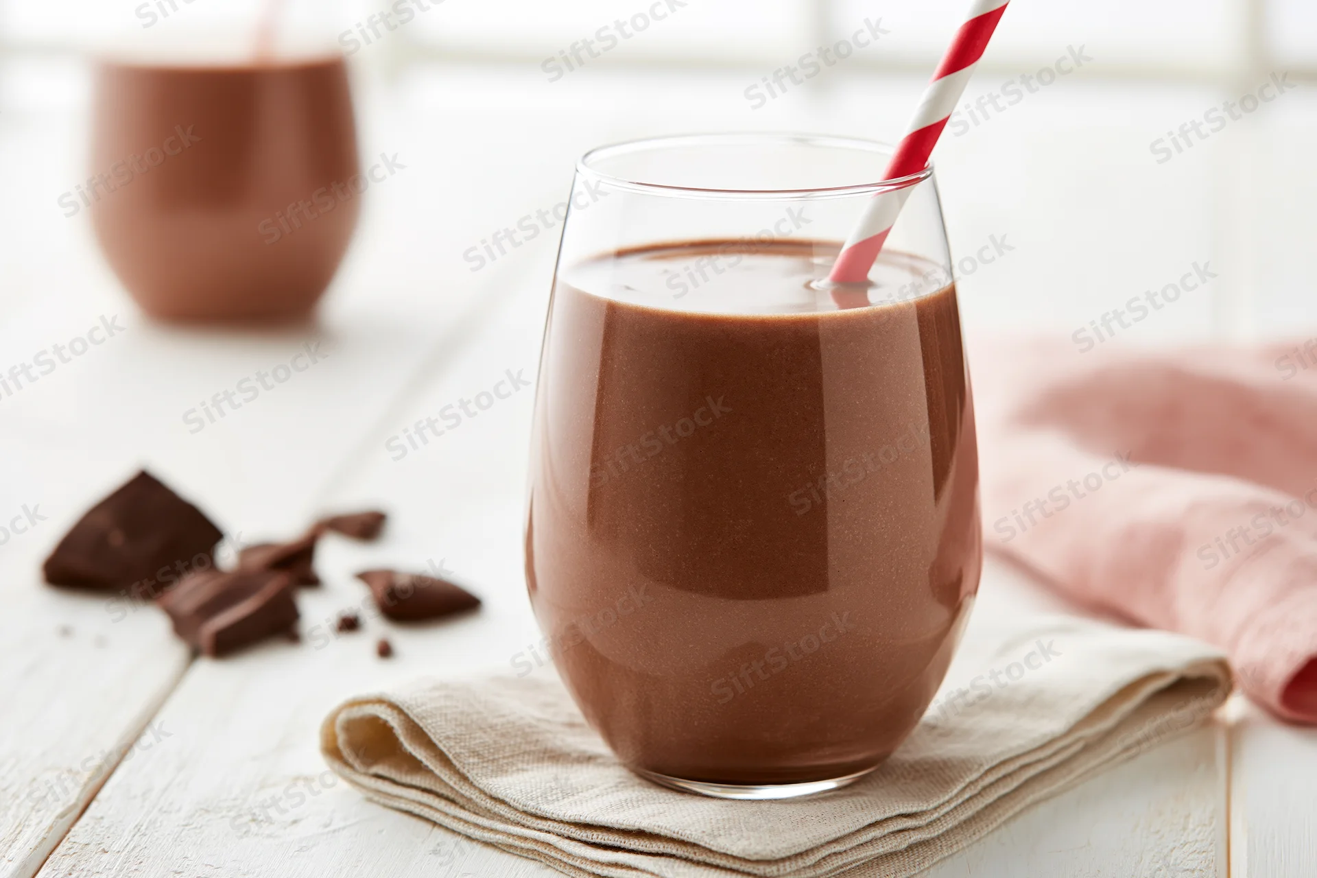 Chocolate milk in a stemless glass with a red-and-white striped straw on a cloth napkin on a white wooden table