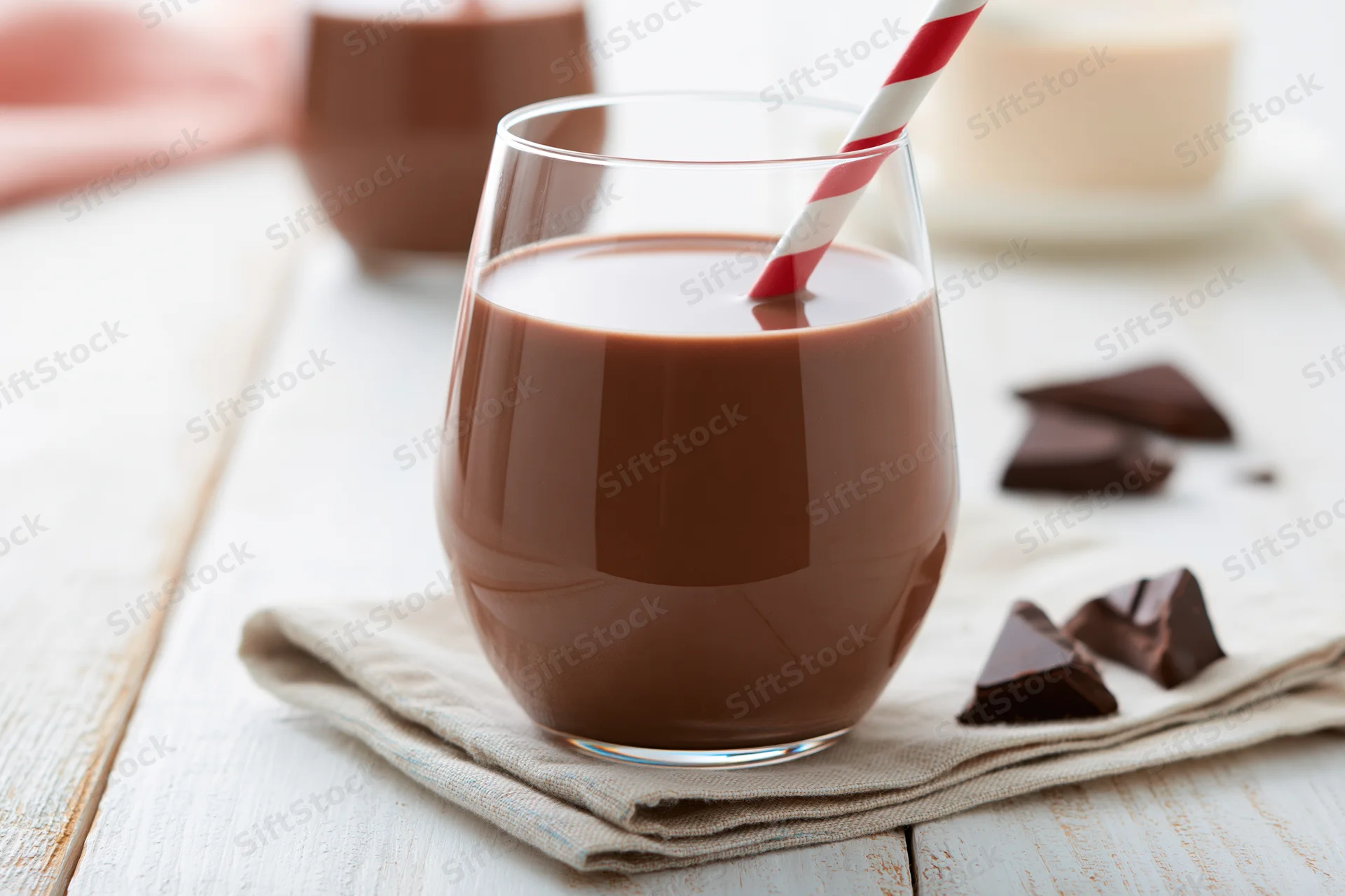 Chocolate milk in a stemless glass with a red and white striped straw on a wooden table with chocolate pieces nearby