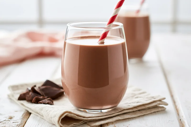 Chocolate milk in a stemless glass with a red-and-white striped straw on a napkin beside chocolate pieces on a white wooden table