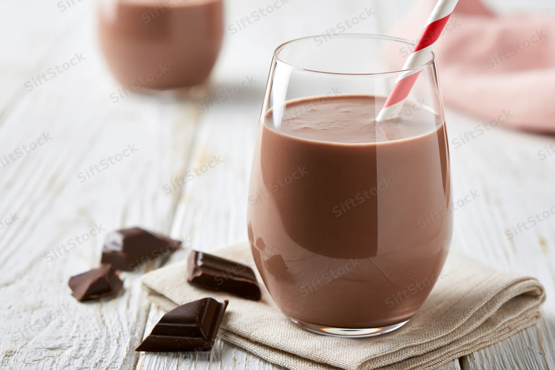 Chocolate milk in a stemless glass with a red-and-white striped straw on a wooden table beside chocolate pieces and a napkin
