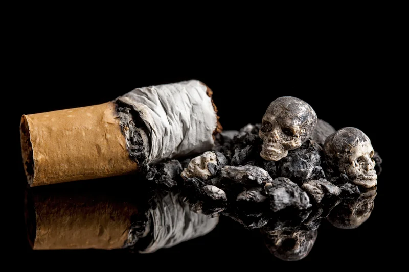 Crushed cigarette butt with ash and miniature skulls on reflective black surface