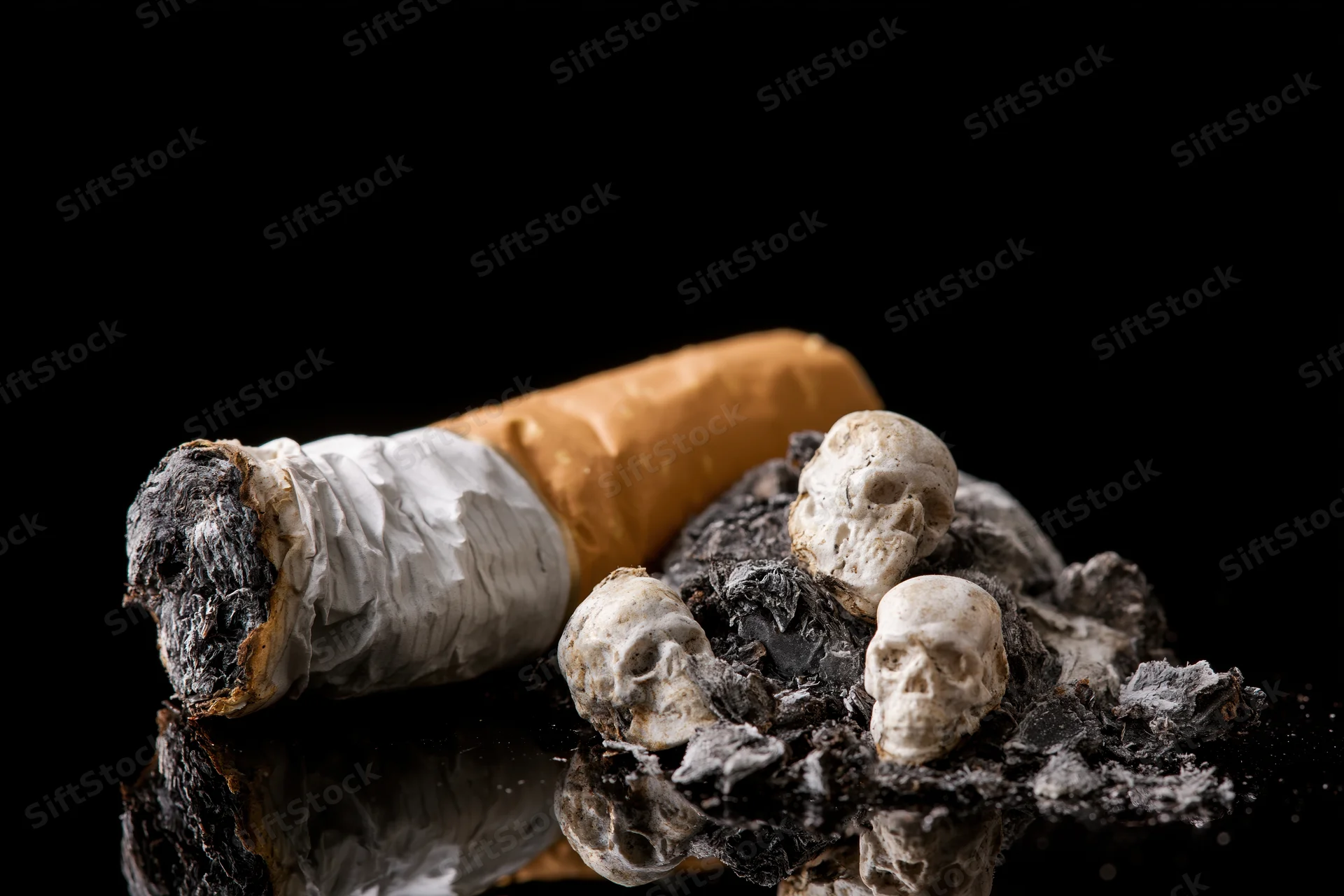 Stubbed-out cigarette resting on ash with miniature skulls on a reflective black surface