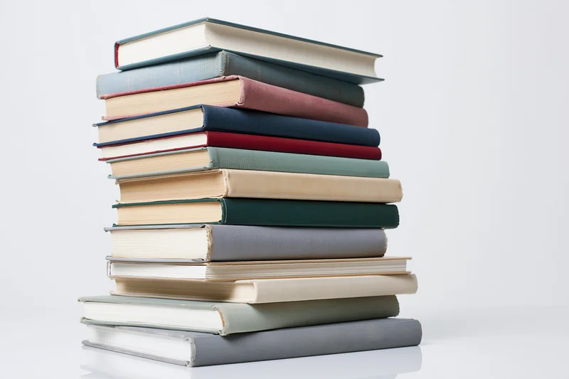 Tall stack of assorted hardcover books arranged vertically on a white surface