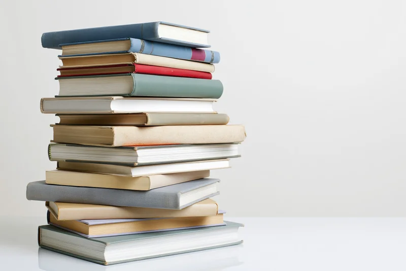 Tall stack of assorted hardcover books arranged vertically on a white surface