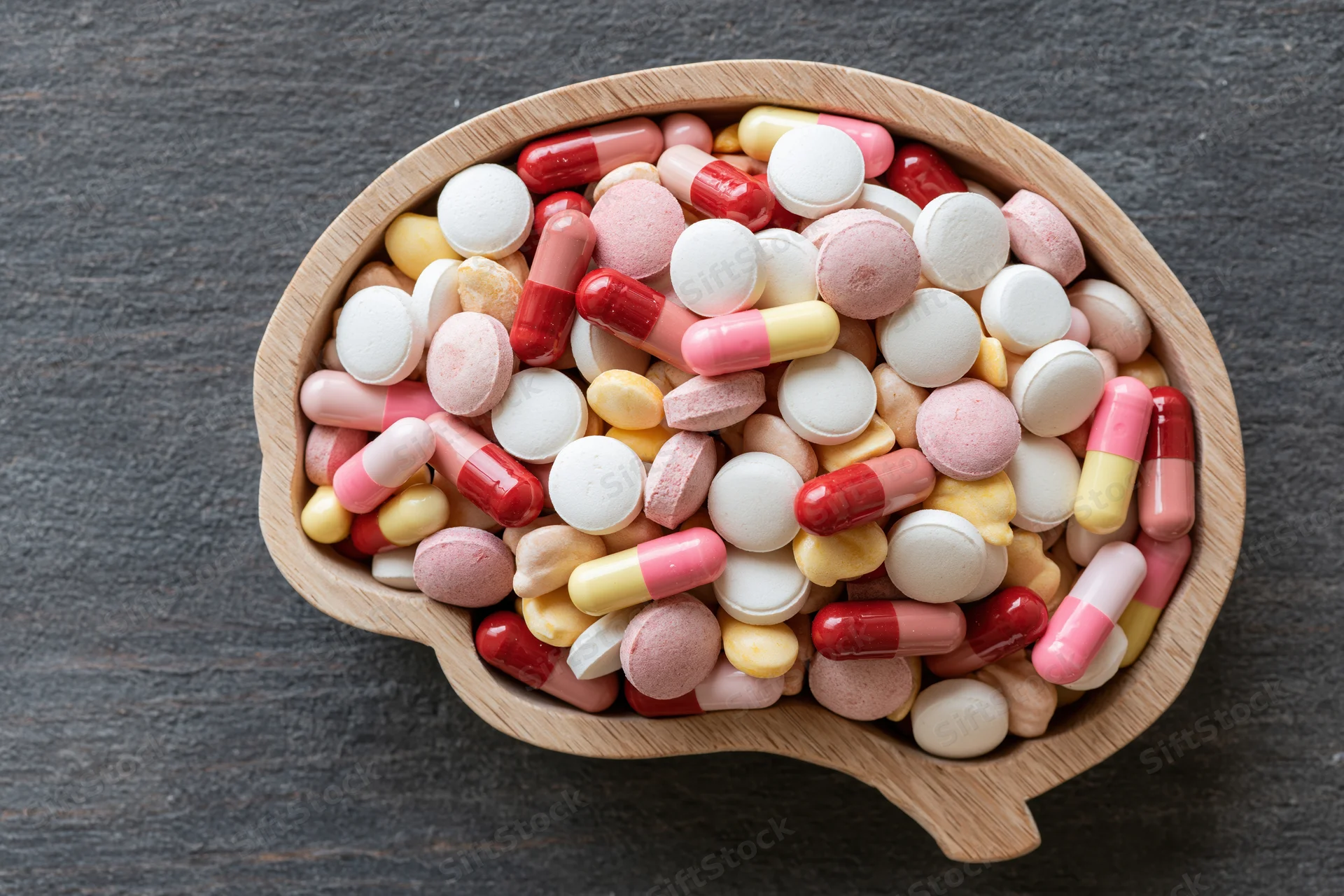 Wooden brain-shaped tray filled with assorted pills and capsules on dark surface