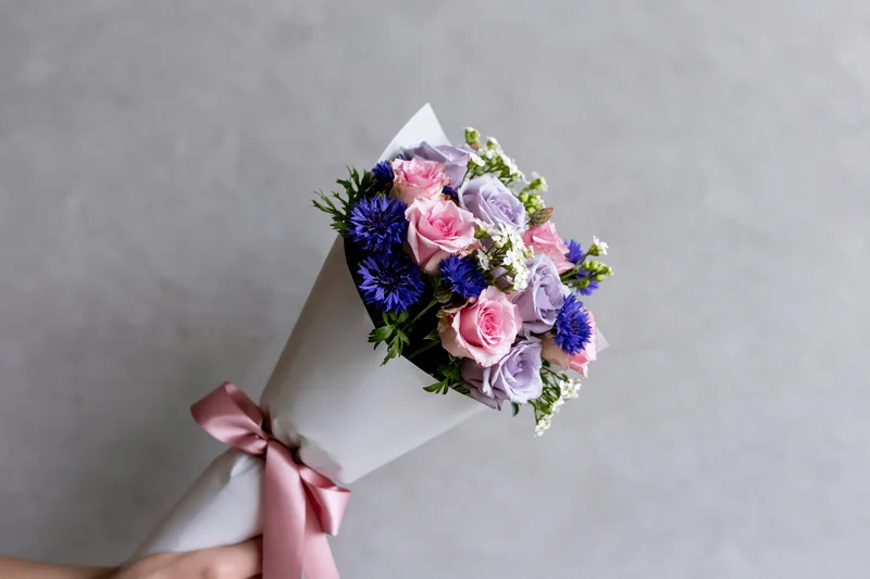 Hand holding a wrapped bouquet of pink and purple roses with blue flowers against a flat gray wall
