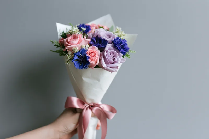 Hand holding a wrapped bouquet of pink and purple roses with blue carnations and greenery against a grey wall