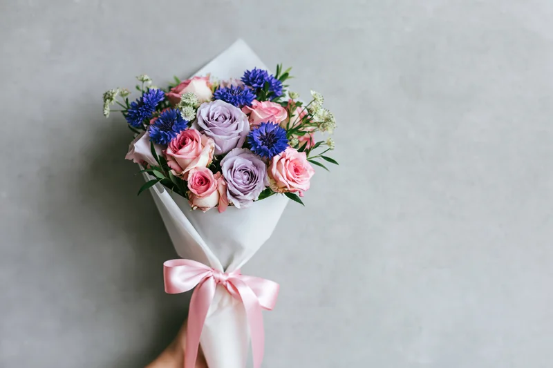 Hand holding a wrapped bouquet of pink, blue and lavender roses with greenery and a pink ribbon against a grey background