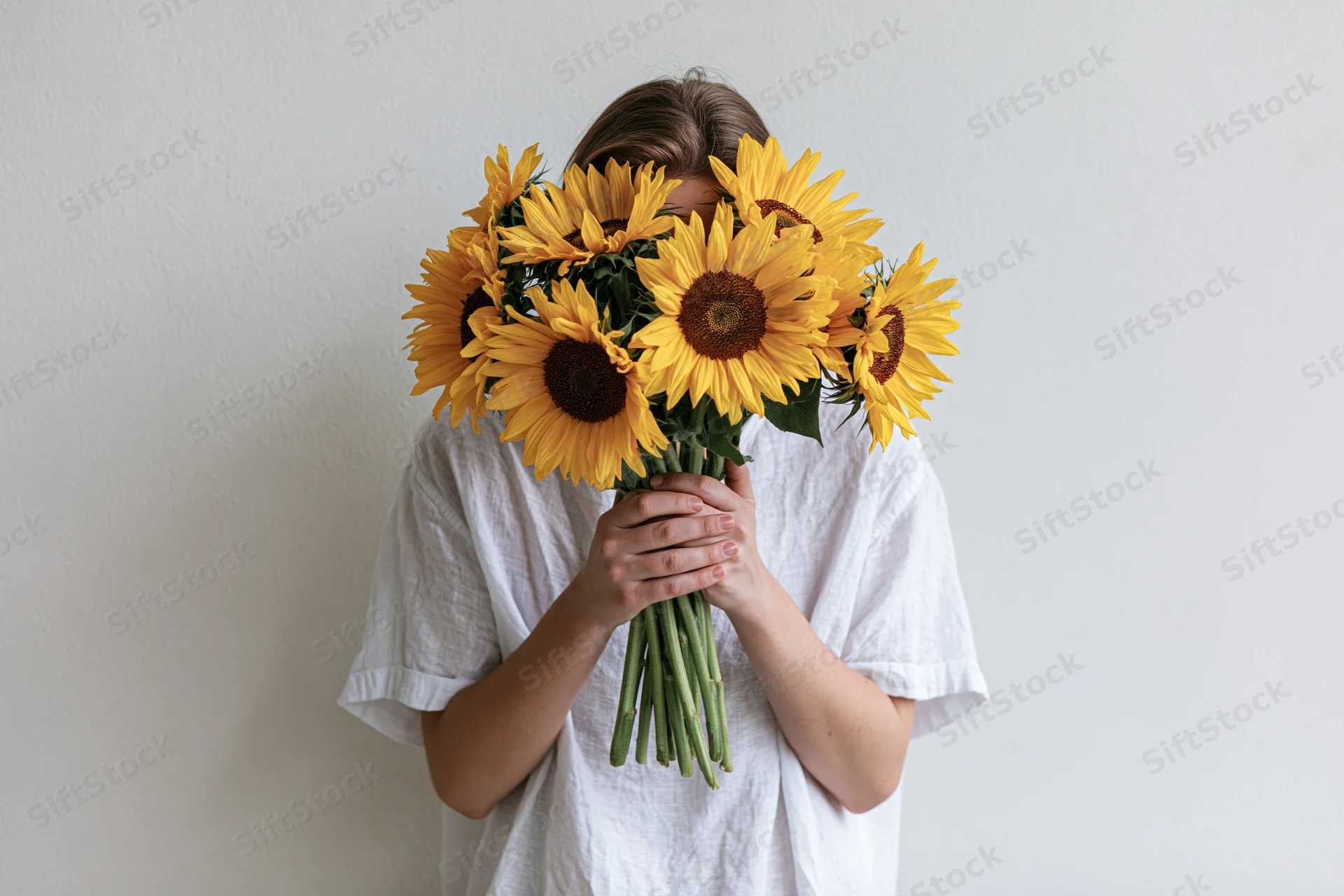 Young woman holding large bouquet of sunflowers in front of her face against white wall