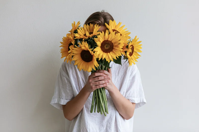 Young woman holding large bouquet of sunflowers in front of her face against white wall