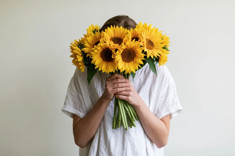 Young woman holding bouquet of sunflowers in front of her face against plain white background