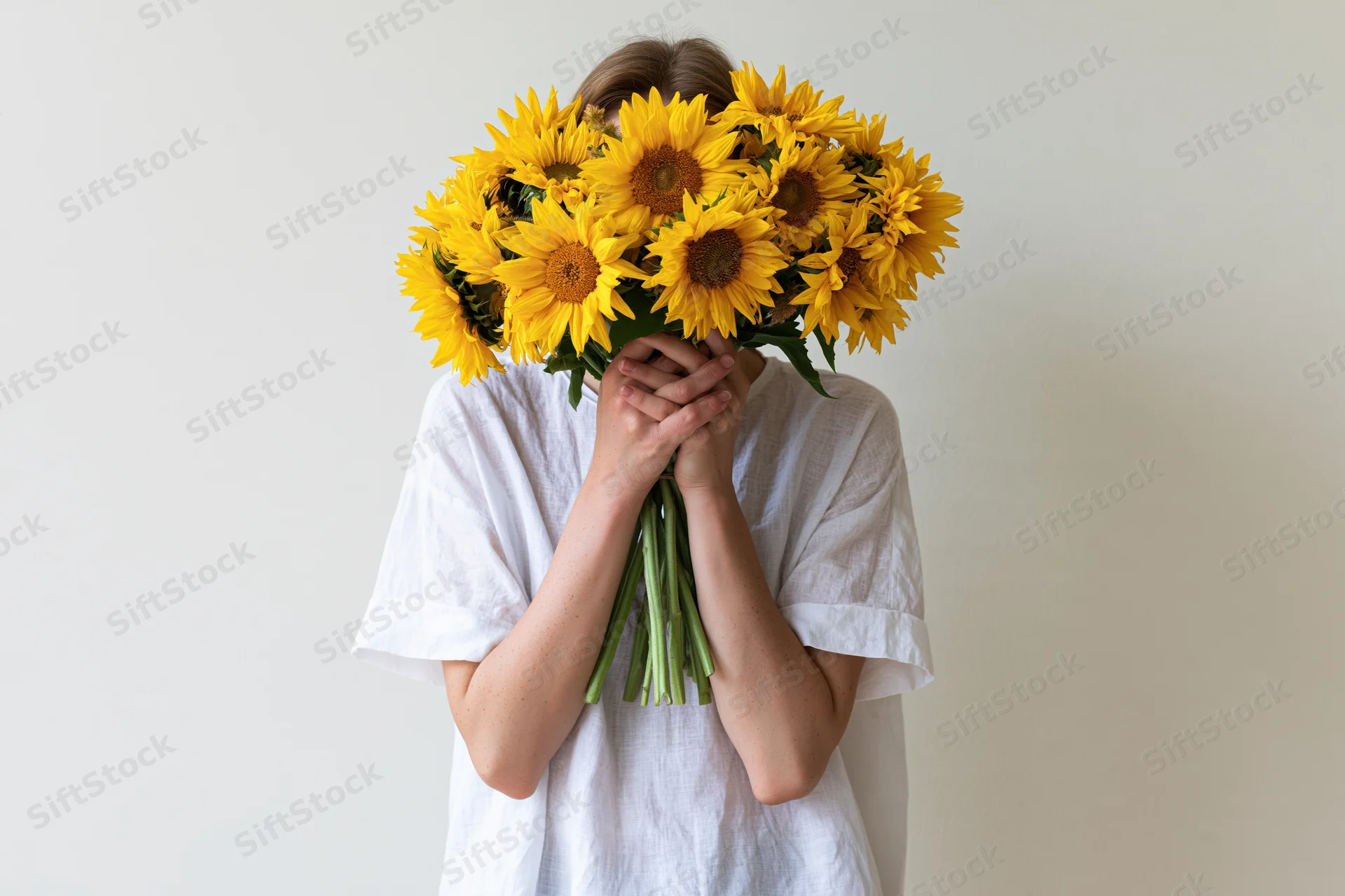 Young woman holding large bouquet of sunflowers in front of her face against plain white background