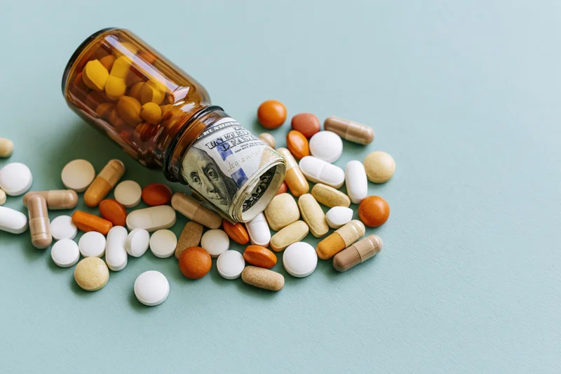 Prescription pill bottle tipped over with scattered pills and a roll of hundred-dollar bills on a light blue surface