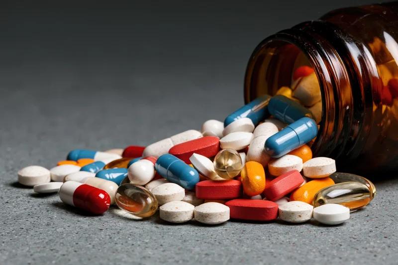 Scattered pills and capsules spilling from an amber glass bottle on a gray surface