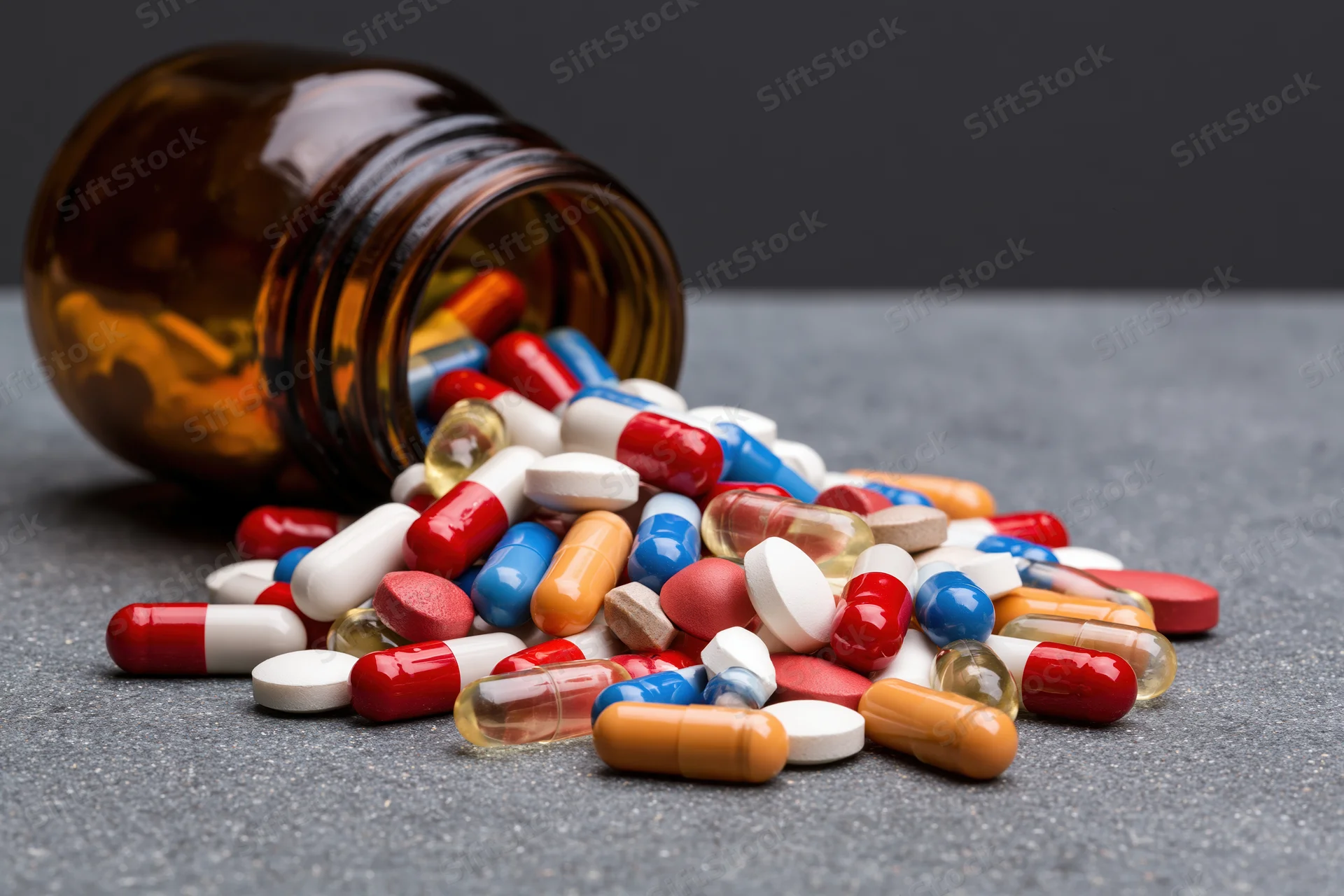 Scattered pills and capsules spilling from an amber glass bottle onto a dark gray surface