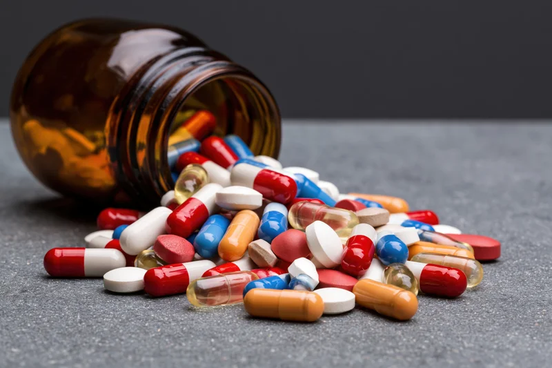 Scattered pills and capsules spilling from an amber glass bottle onto a dark gray surface