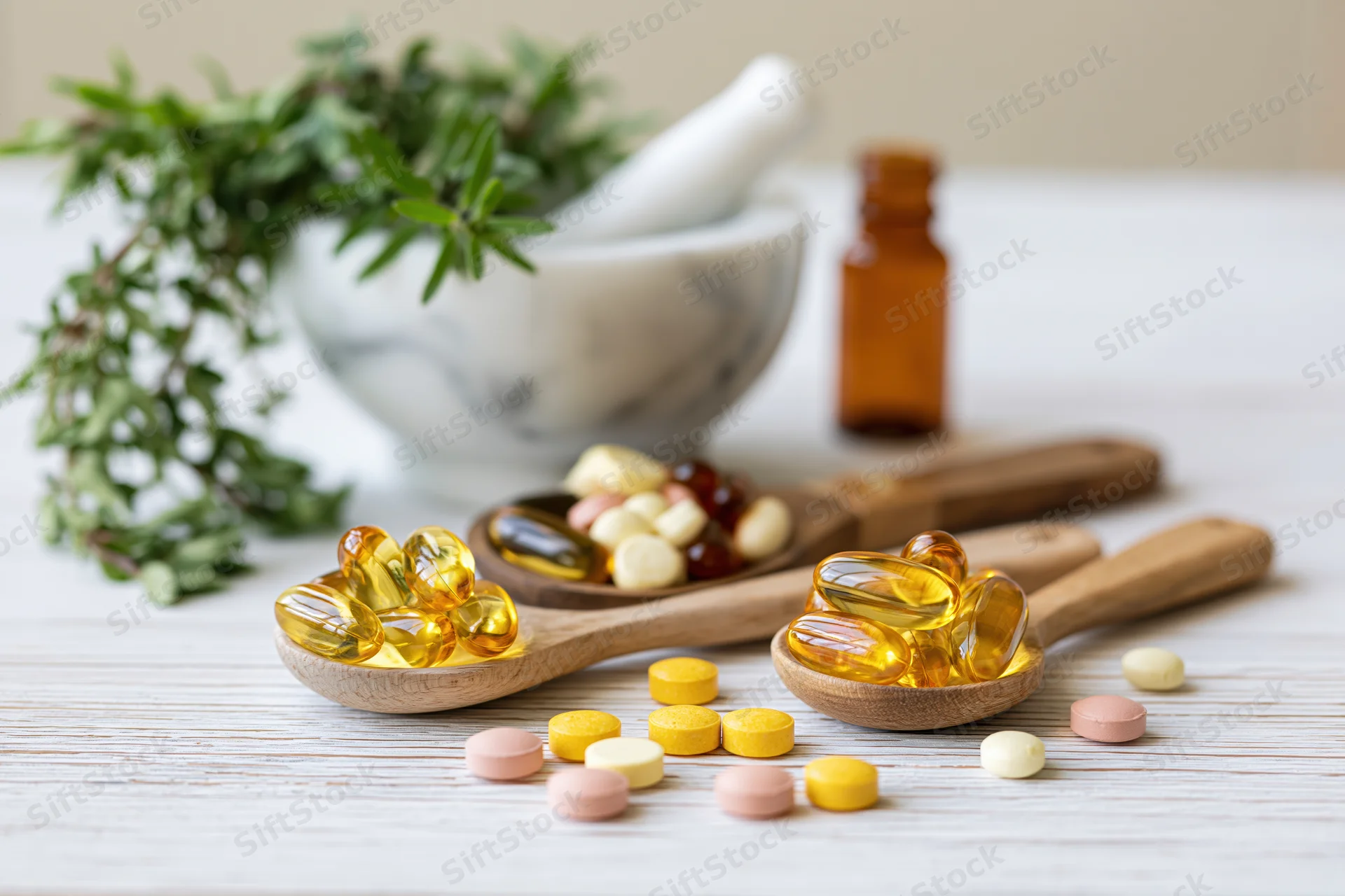 Herbal supplement capsules and gel caps arranged on wooden spoons with herbs and mortar on a light wood surface