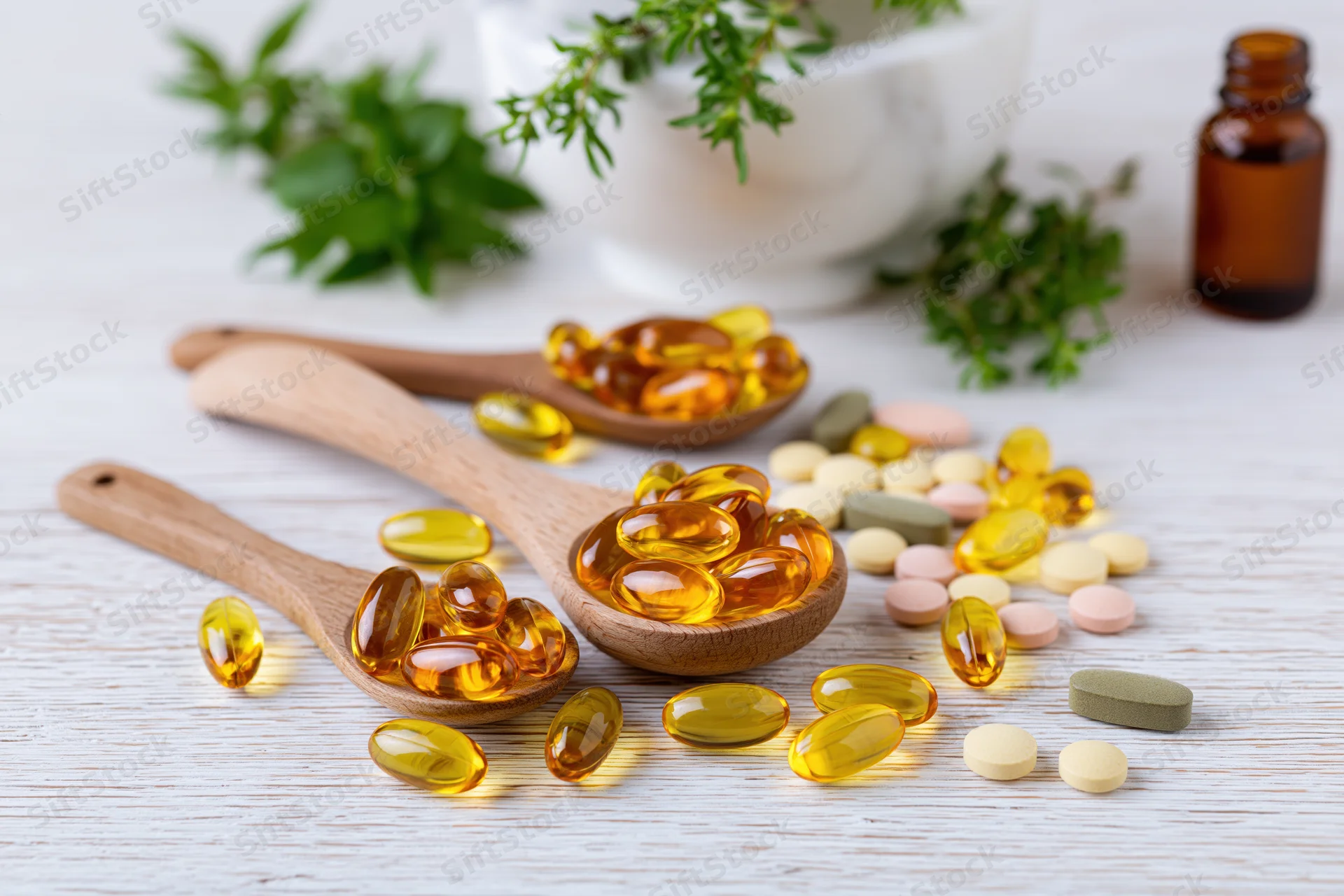 Herbal supplement capsules and gel caps in wooden spoons on a light wooden surface with fresh herbs and a mortar in the background
