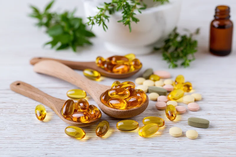 Herbal supplement capsules and gel caps in wooden spoons on a light wooden surface with fresh herbs and a mortar in the background