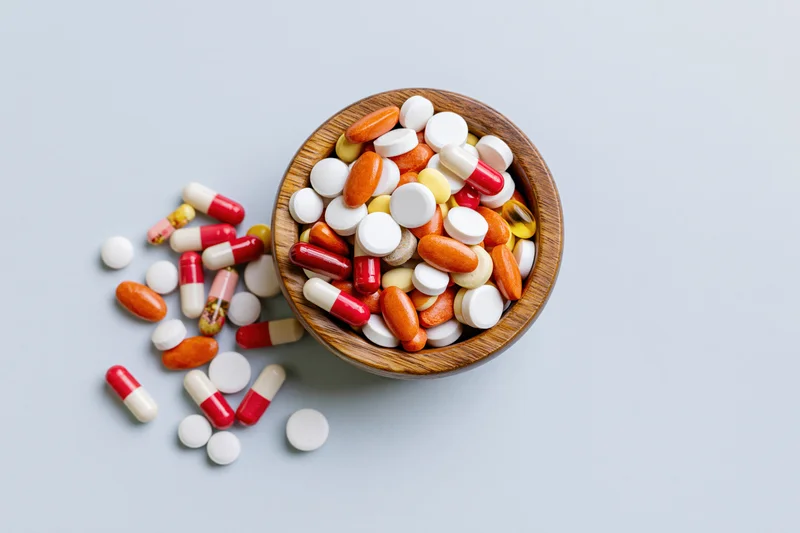 Assorted pharmaceutical pills and capsules overflowing from a wooden bowl onto a light gray surface